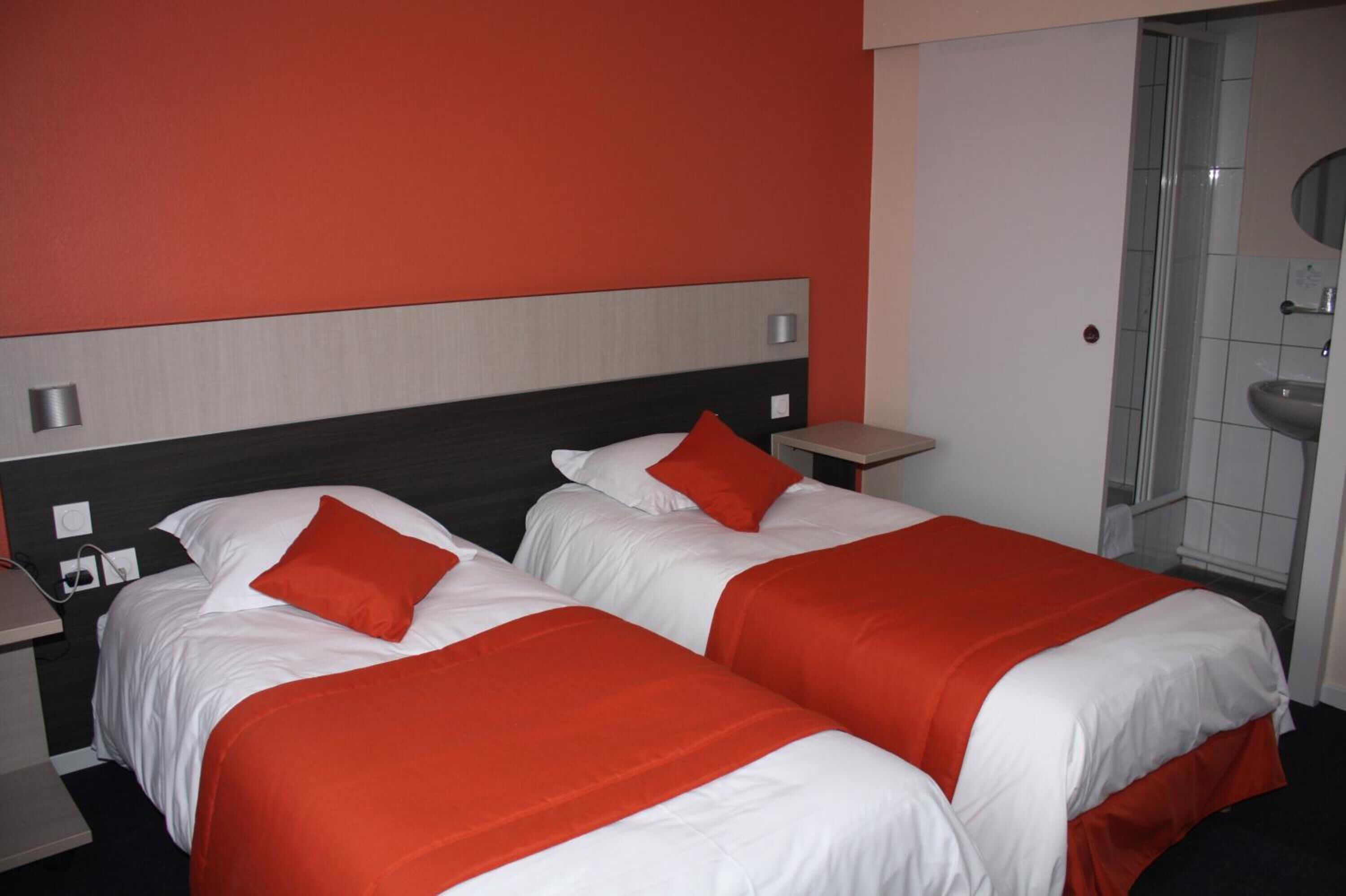 A hotel room with two beds, each with white bedding and red accents, a grey headboard, a small bedside table, and a bathroom with a sink and mirror.