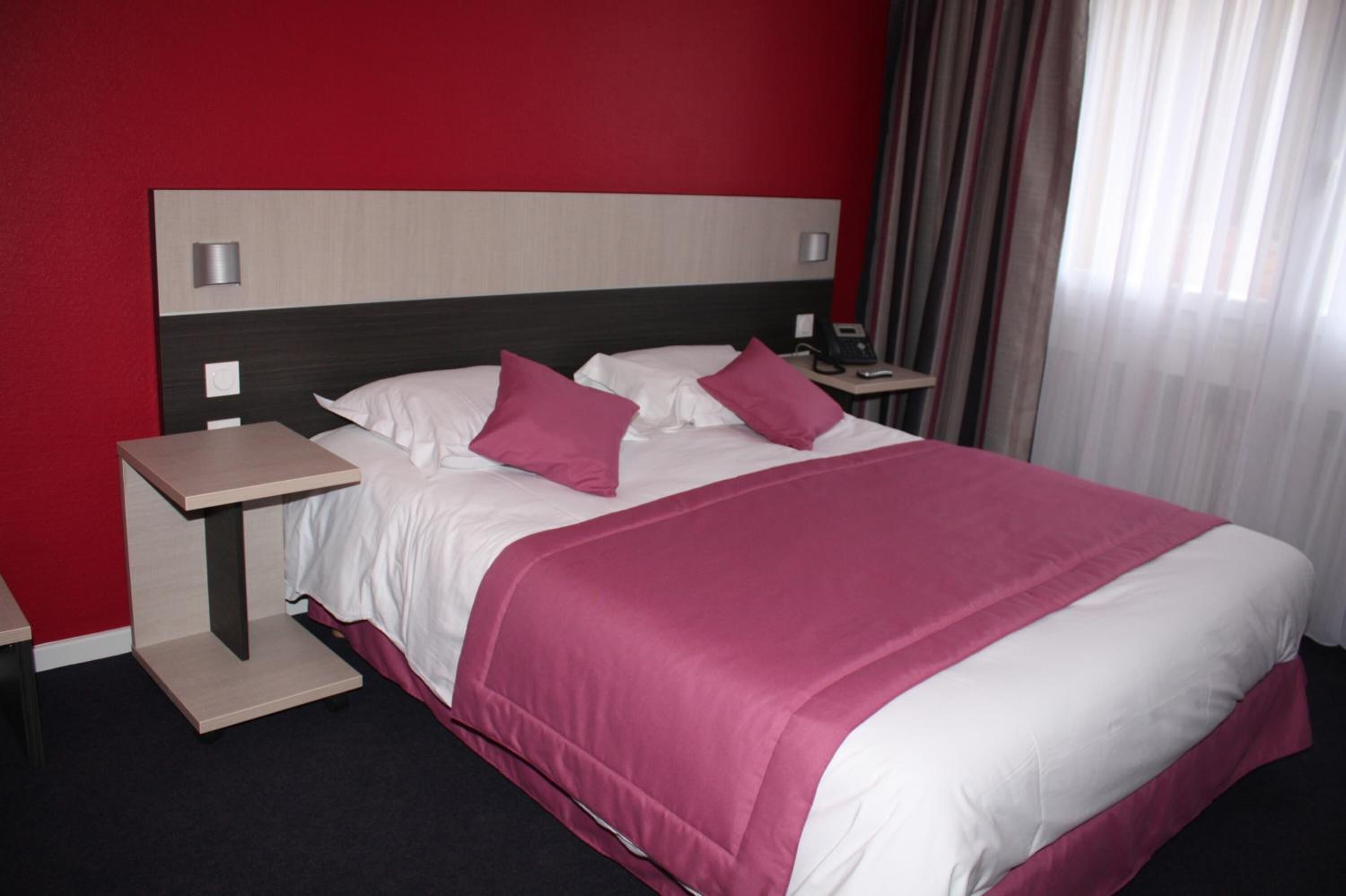 Quarto de hotel com uma cama, mesa de cabeceira e uma parede vermelha.