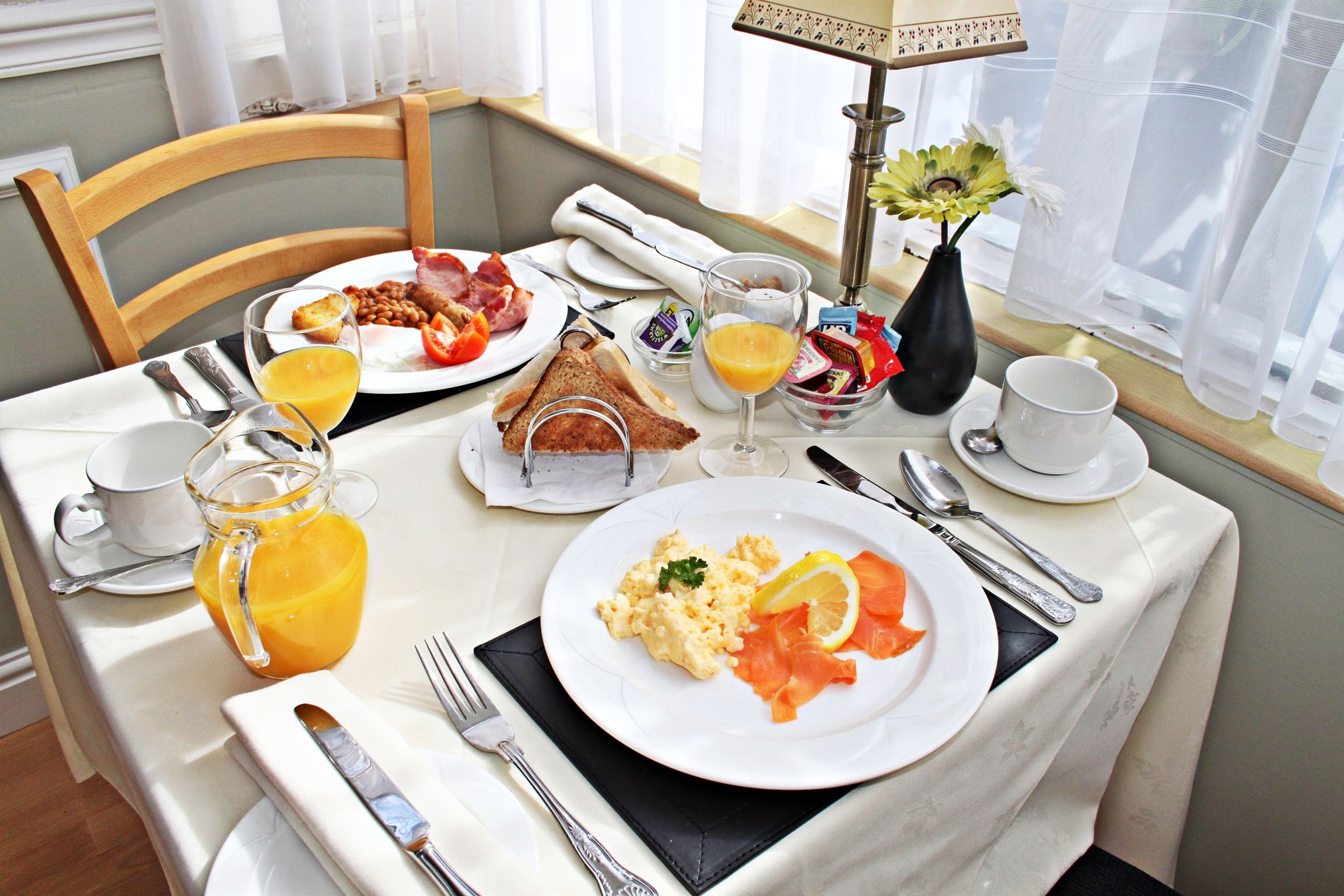 A breakfast setting with a table set for two, including plates of eggs, bacon, toast, and a glass of orange juice.