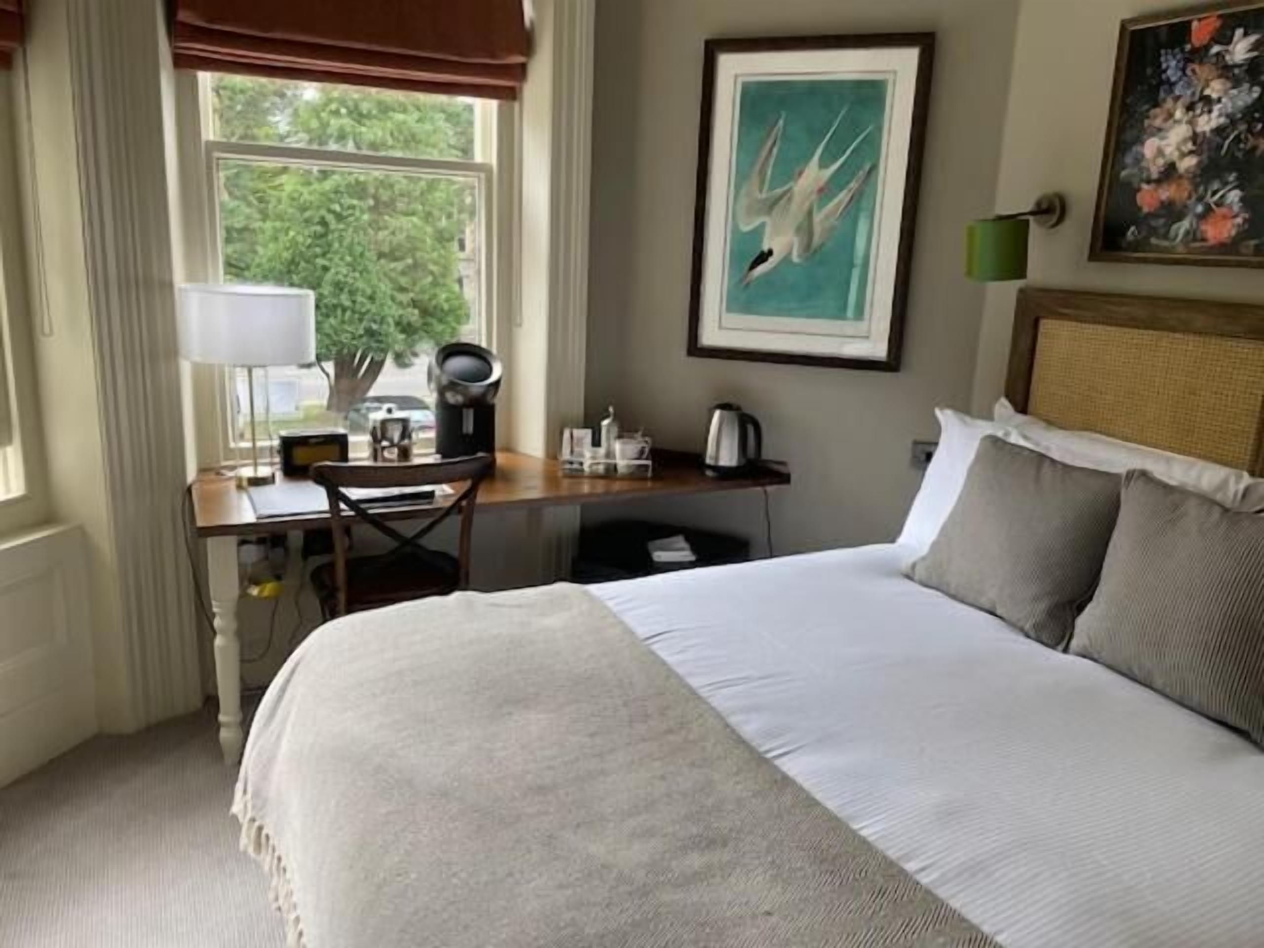 A hotel room with a bed, a desk with a chair, a window with curtains, a painting on the wall, and a lamp.