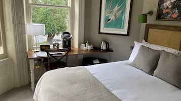 A hotel room with a bed, a desk with a chair, a window with curtains, a painting on the wall, and a lamp.