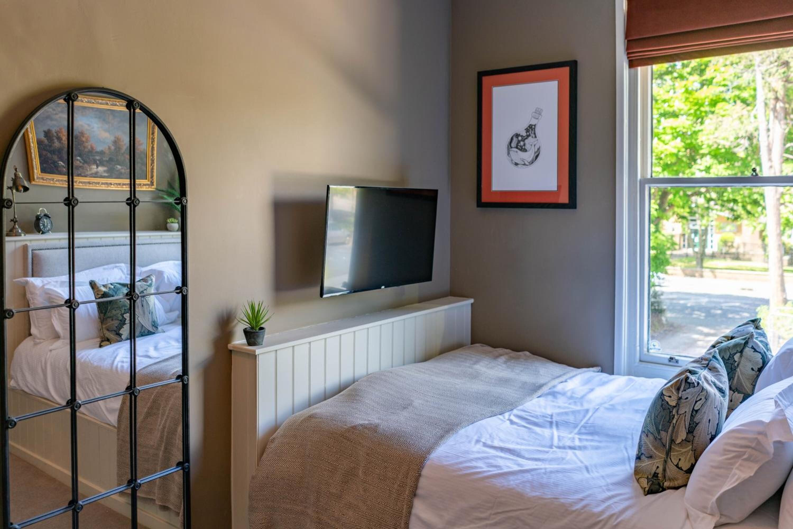 Un dormitorio con cama, televisor montado en la pared, ventana con vistas a árboles y un cuadro enmarcado en la pared.