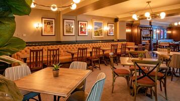 Ein Restaurant mit Sitzecken, Tischen und Stühlen. An der Wand hängen gerahmte Bilder und im Hintergrund ist eine Bar zu sehen.