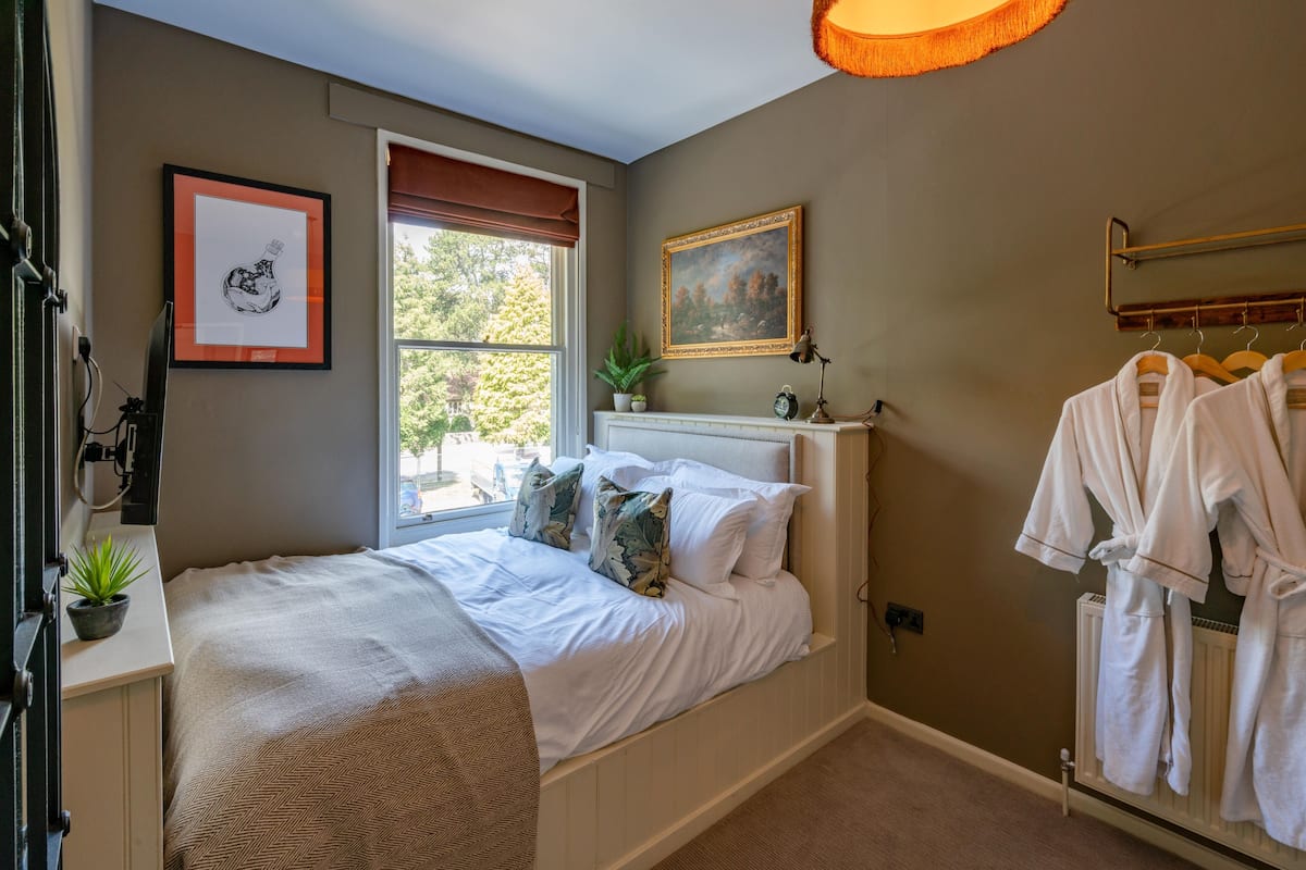 A bedroom with a bed, a window with a view, a painting, and a radiator with robes.