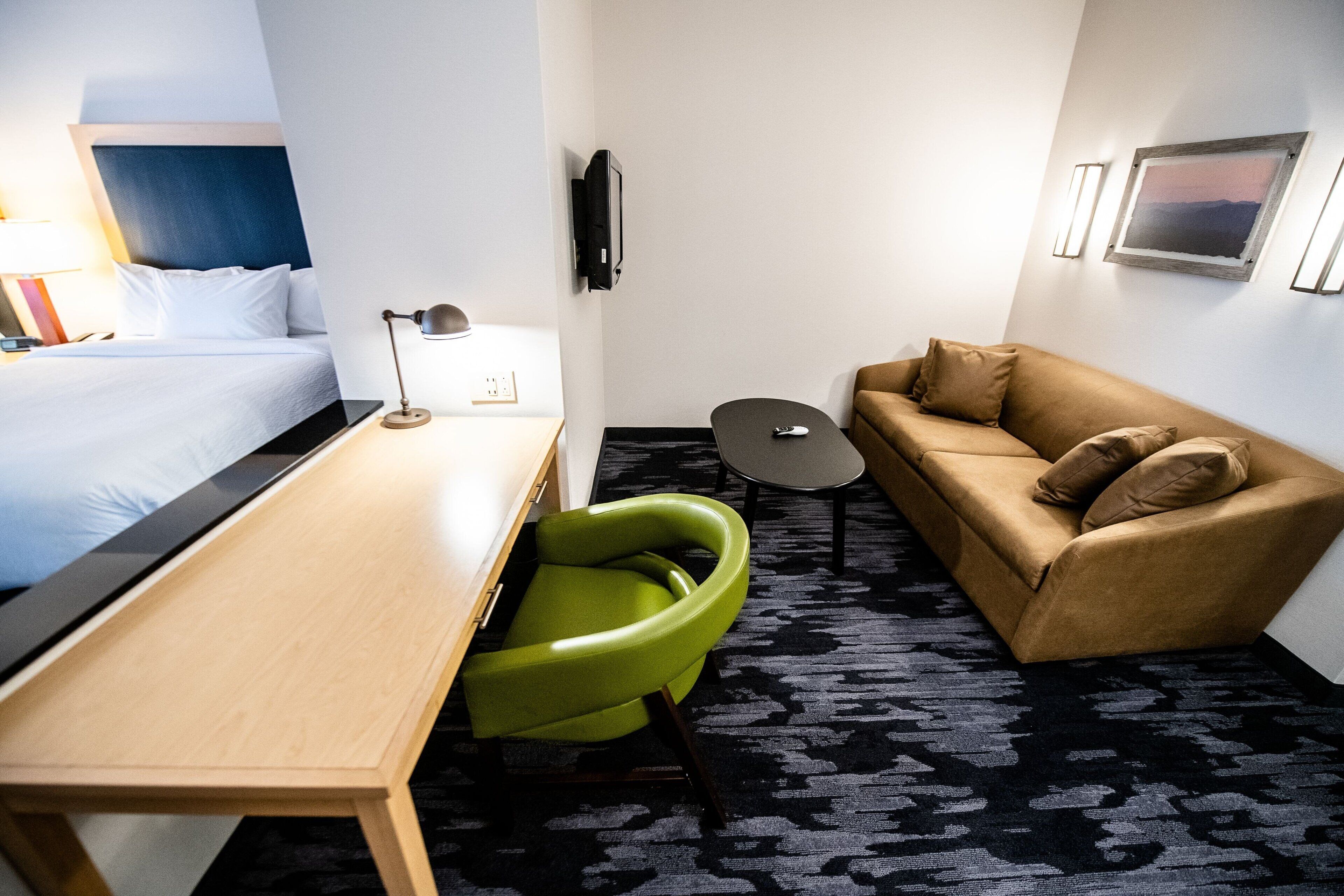 A hotel room with a bed, a desk, a green chair, a sofa, a TV, and a small table.