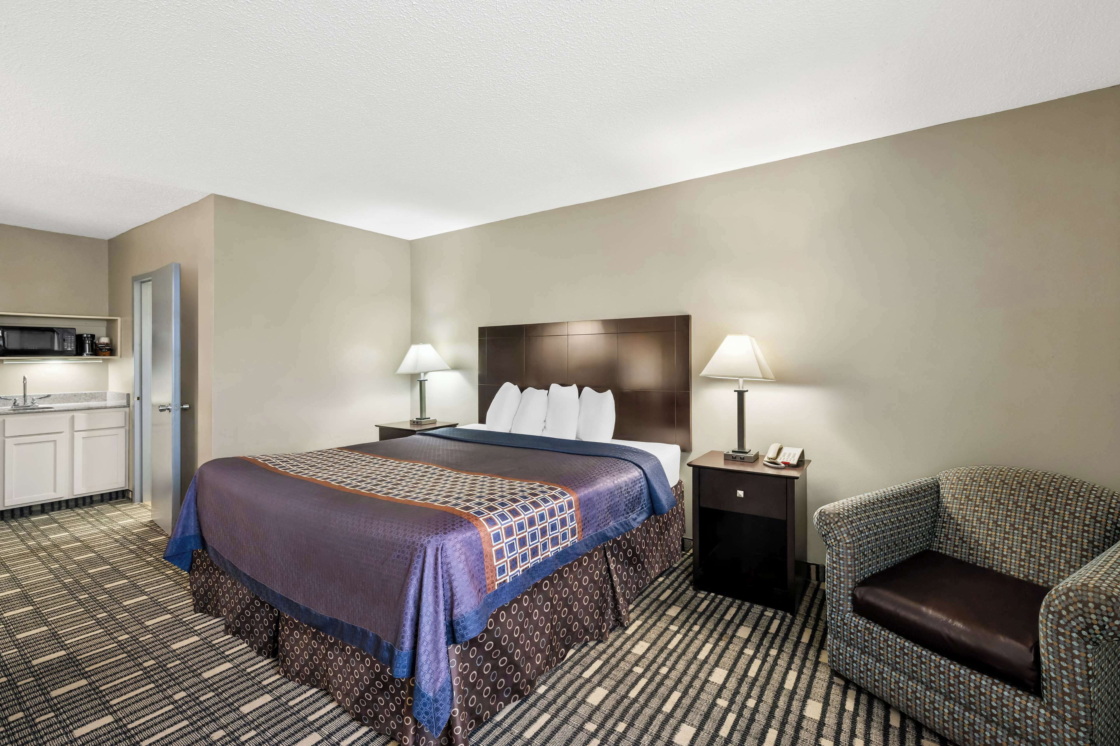 Habitación de hotel con cama, dos lámparas de noche, una silla, mesita de noche y kitchenette con microondas.
