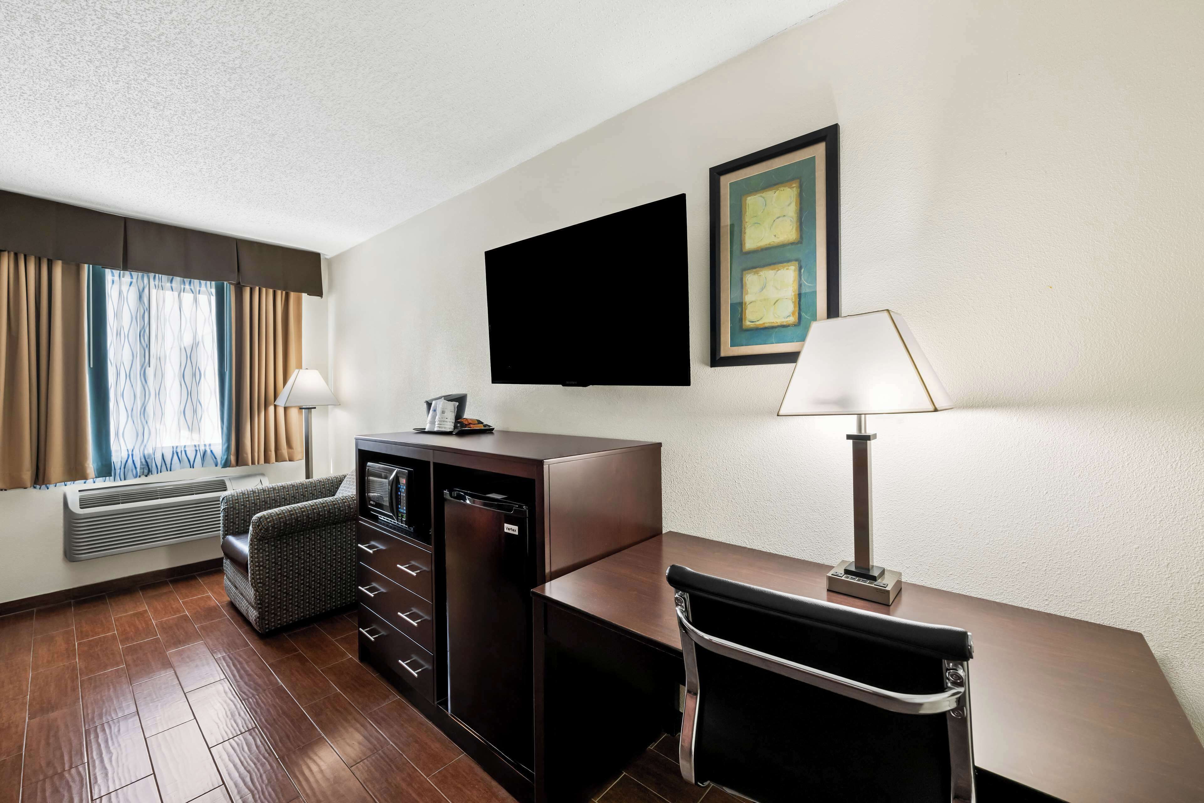 A hotel room with a flat-screen TV, a desk with a lamp, a sofa, a mini-fridge, and a heater.