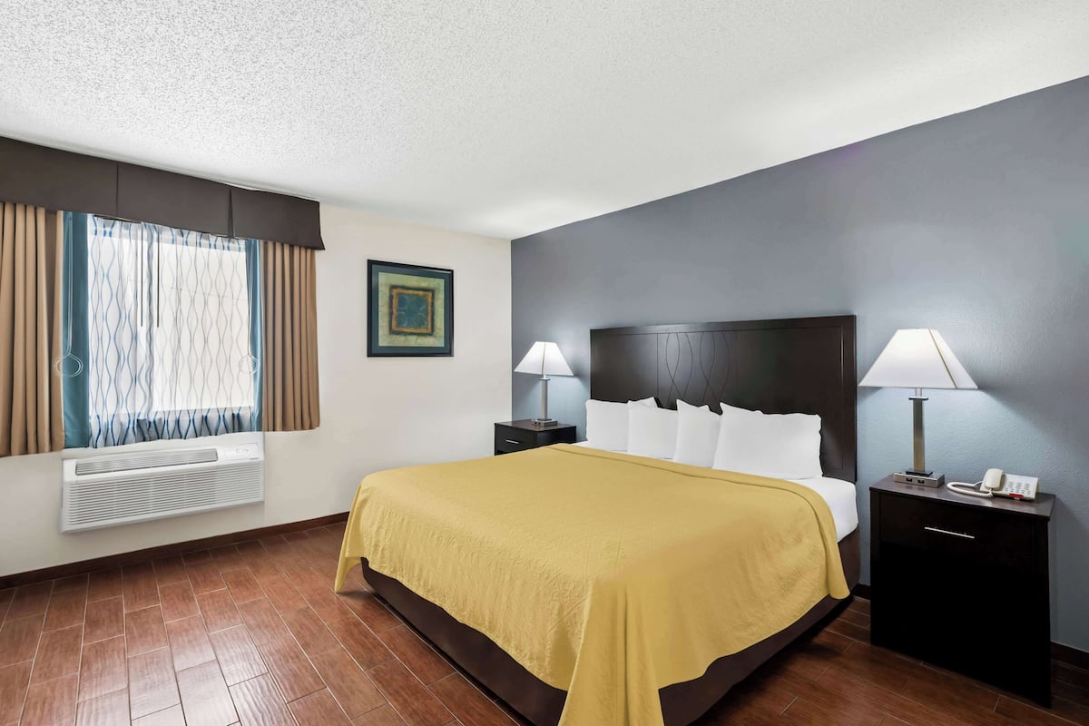 Une chambre d’hôtel comprenant un lit, deux tables de chevet avec des lampes, un climatiseur fixé au mur, une fenêtre avec des rideaux et un tableau encadré accroché au mur.