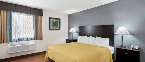A hotel room with a bed, two bedside tables with lamps, a wall-mounted air conditioner, a window with curtains, and a framed picture on the wall.