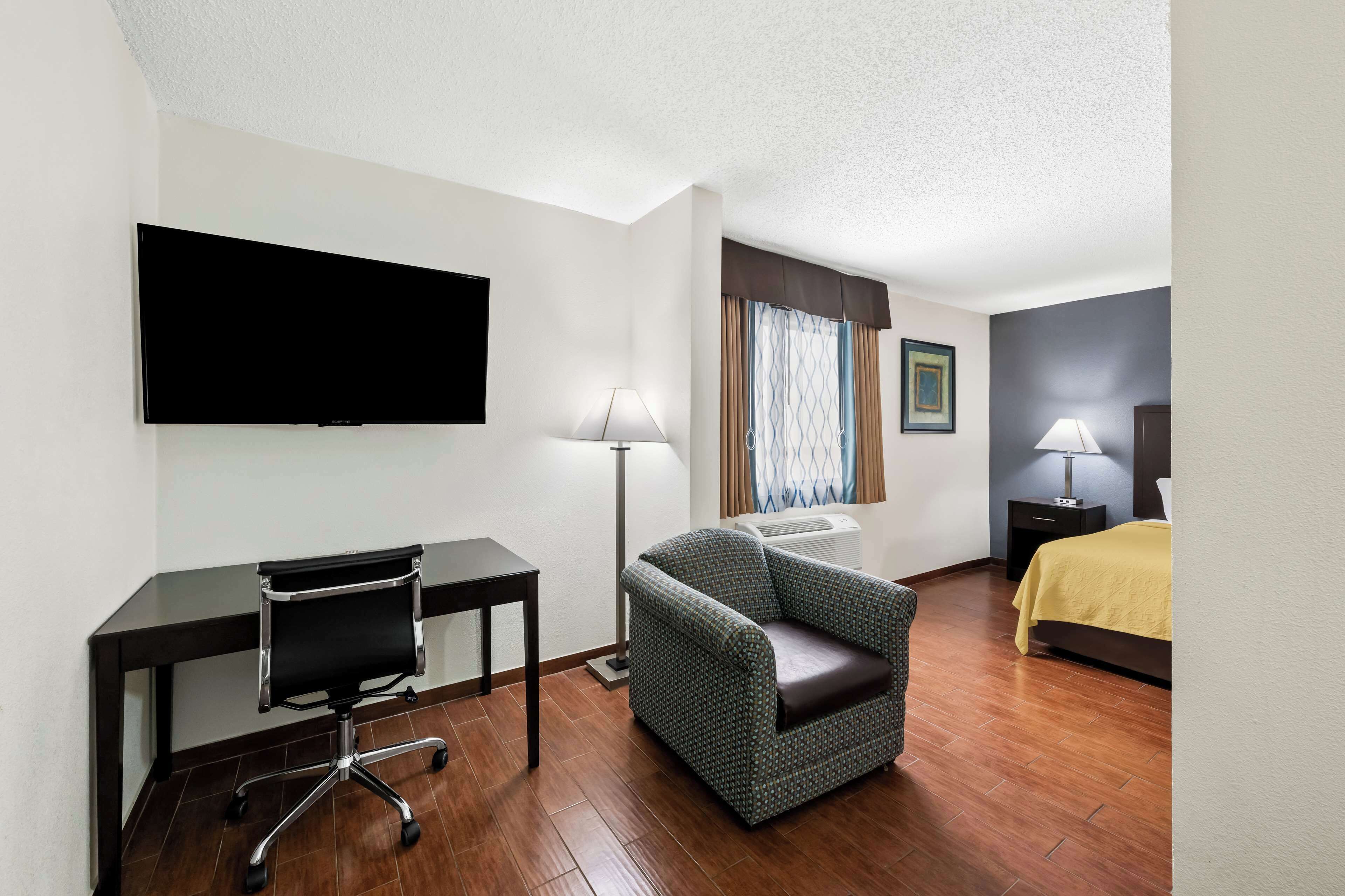 Une chambre d’hôtel équipée d’une télévision à écran plat, d’un bureau avec une chaise, d’un canapé, d’un lit recouvert d’une couvre-lit jaune et de deux lampes.