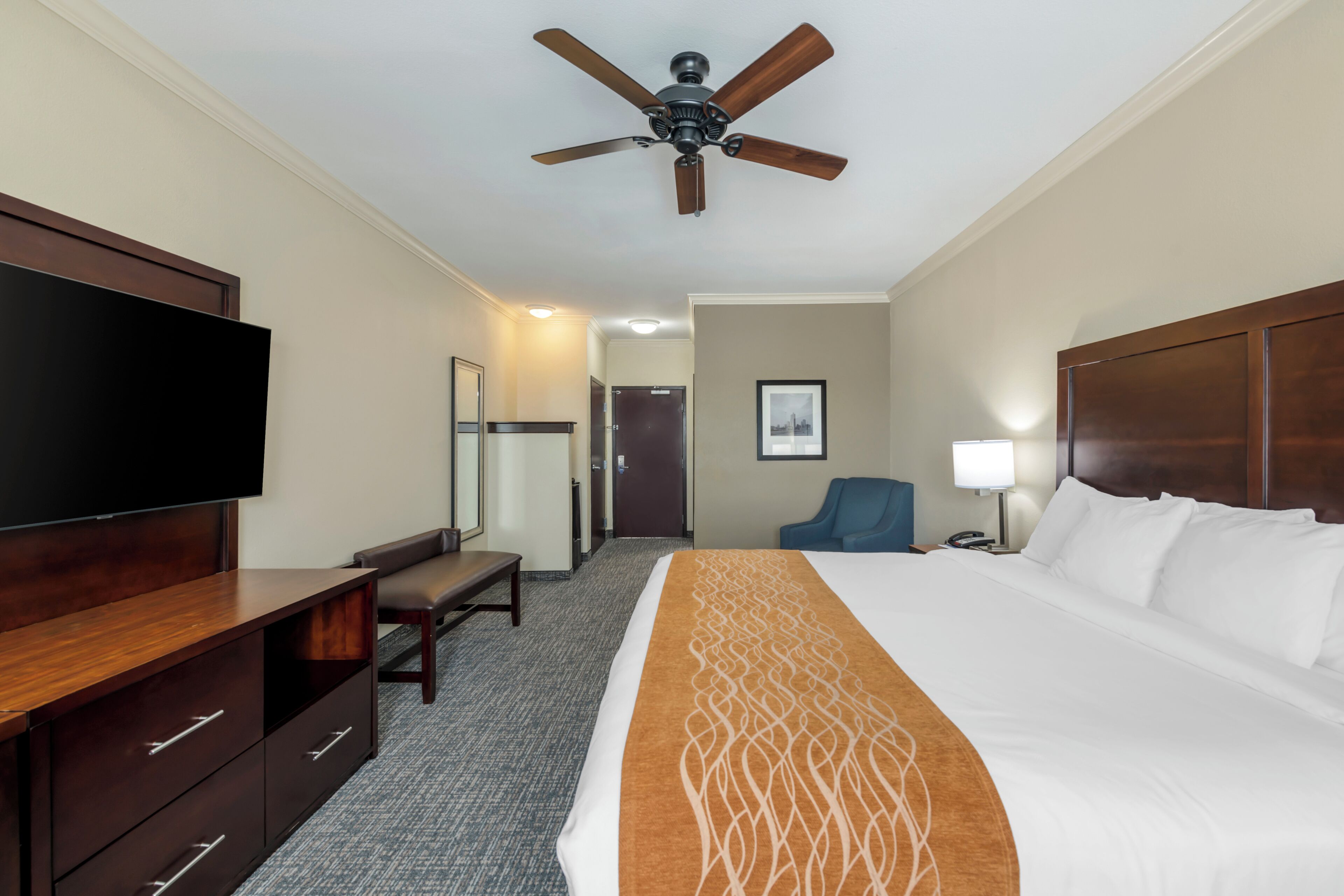 A hotel room with a large bed, a flat-screen TV, a desk with drawers, a chair, a ceiling fan, and a framed picture on the wall.