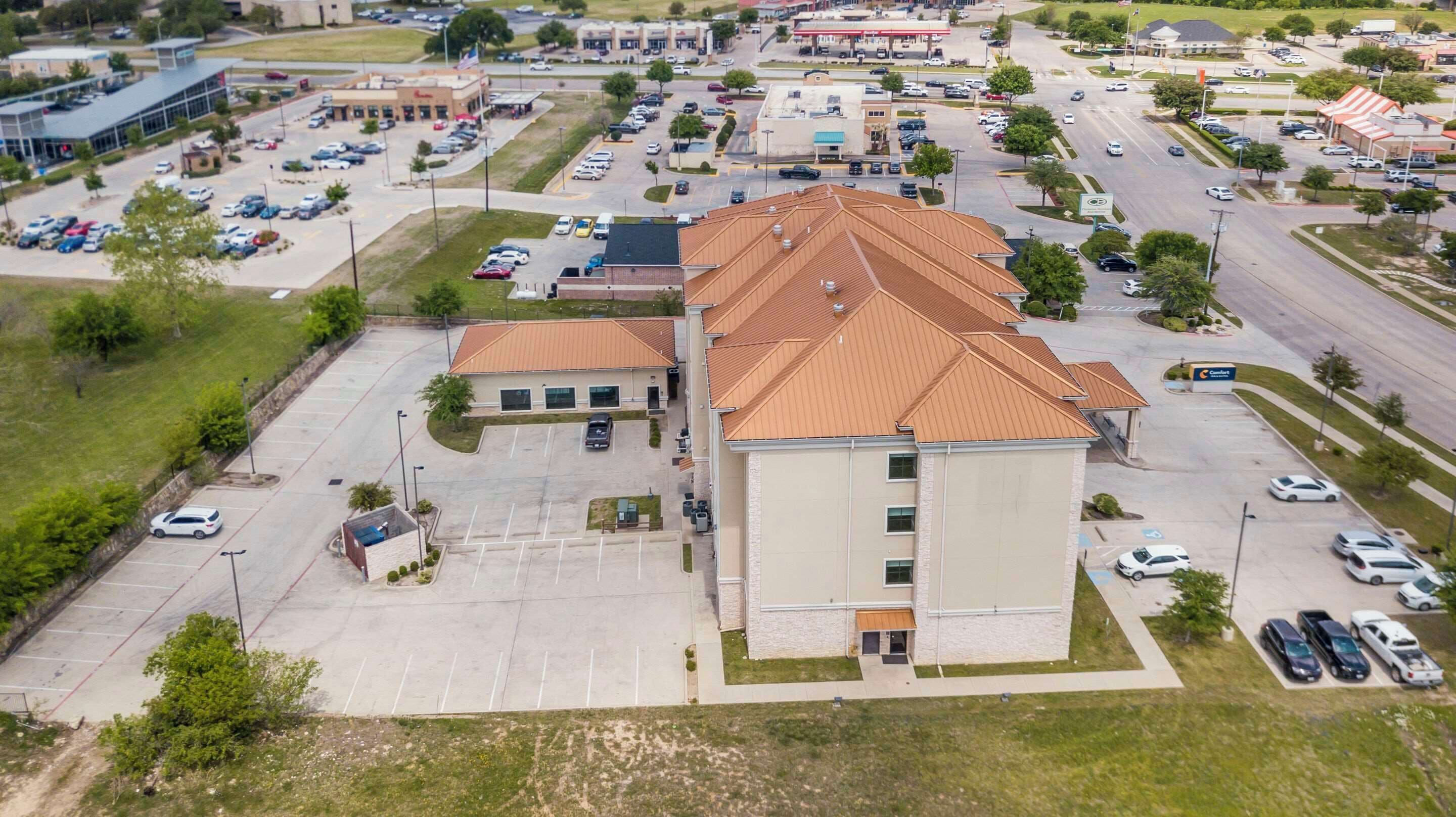 An aerial view of a commercial area with a large building with a brown roof, multiple parking lots, and various businesses including a gas station.