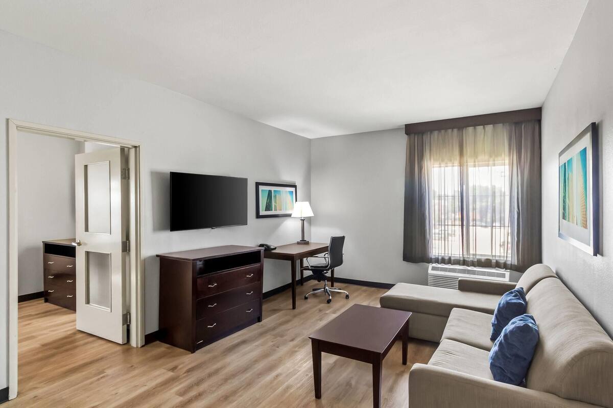 A hotel room with a wooden floor, a desk with a chair, a sofa, a TV mounted on the wall, and a window with curtains.