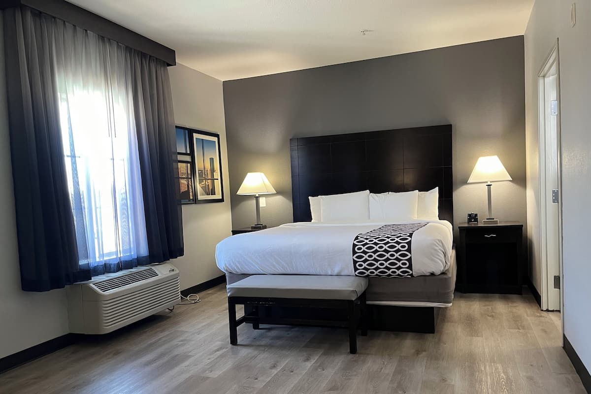 A hotel room with a large bed, two bedside tables with lamps, a window with curtains, and a wall-mounted air conditioner.