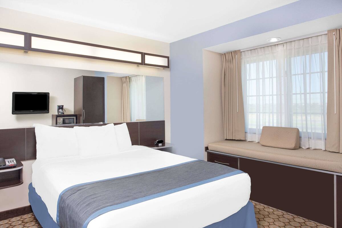 A hotel room with a bed, a TV, a microwave, a bedside table, and a window with curtains.