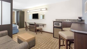 Habitación de hotel compacta con kitchenette, cama con ropa de cama azul, pequeña mesa de comedor con sillas y un televisor montado en la pared.