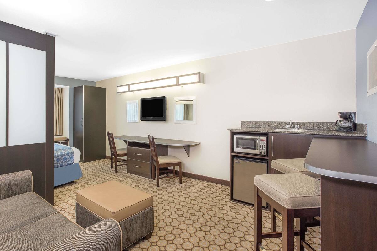 A compact hotel room with a kitchenette, a bed with blue bedding, a small dining table with chairs, and a TV mounted on the wall.