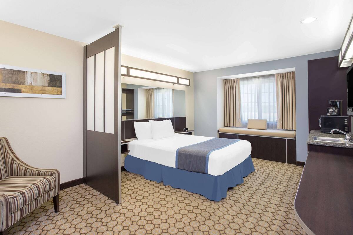 A hotel room with a bed, a chair, a desk with a sink, and a window with curtains.