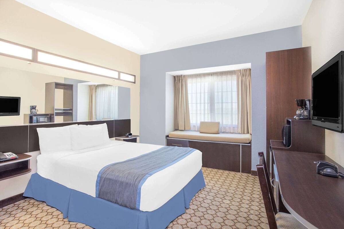 A hotel room with a bed, a desk with a chair, a television, a window with curtains, and a coffee machine.