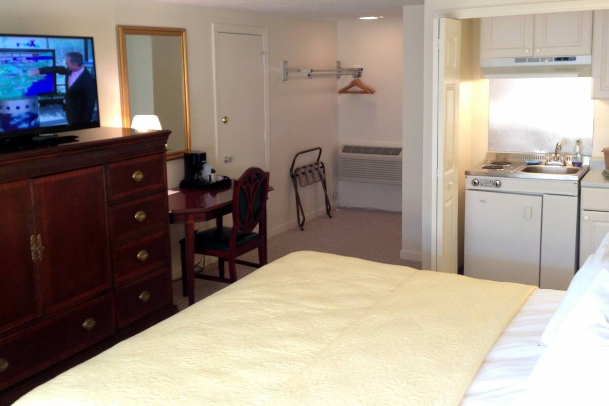 Une chambre d’hôtel équipée d’un lit, d’un bureau, d’une télévision, d’un miroir, d’une kitchenette et d’un porte-manteau mural.