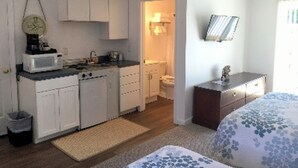 A compact kitchen with white cabinets, a microwave, and a sink. There is a small rug on the floor and a bed with a floral bedspread in the adjacent room. - Craigville Beach Inn (Centerville)