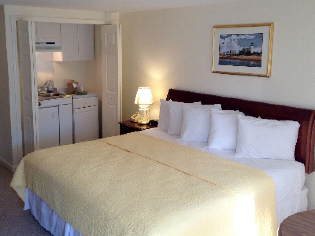 Habitación de hotel con una cama grande, una mesita de noche con lámpara, un cuadro en la pared y una kitchenette al fondo.