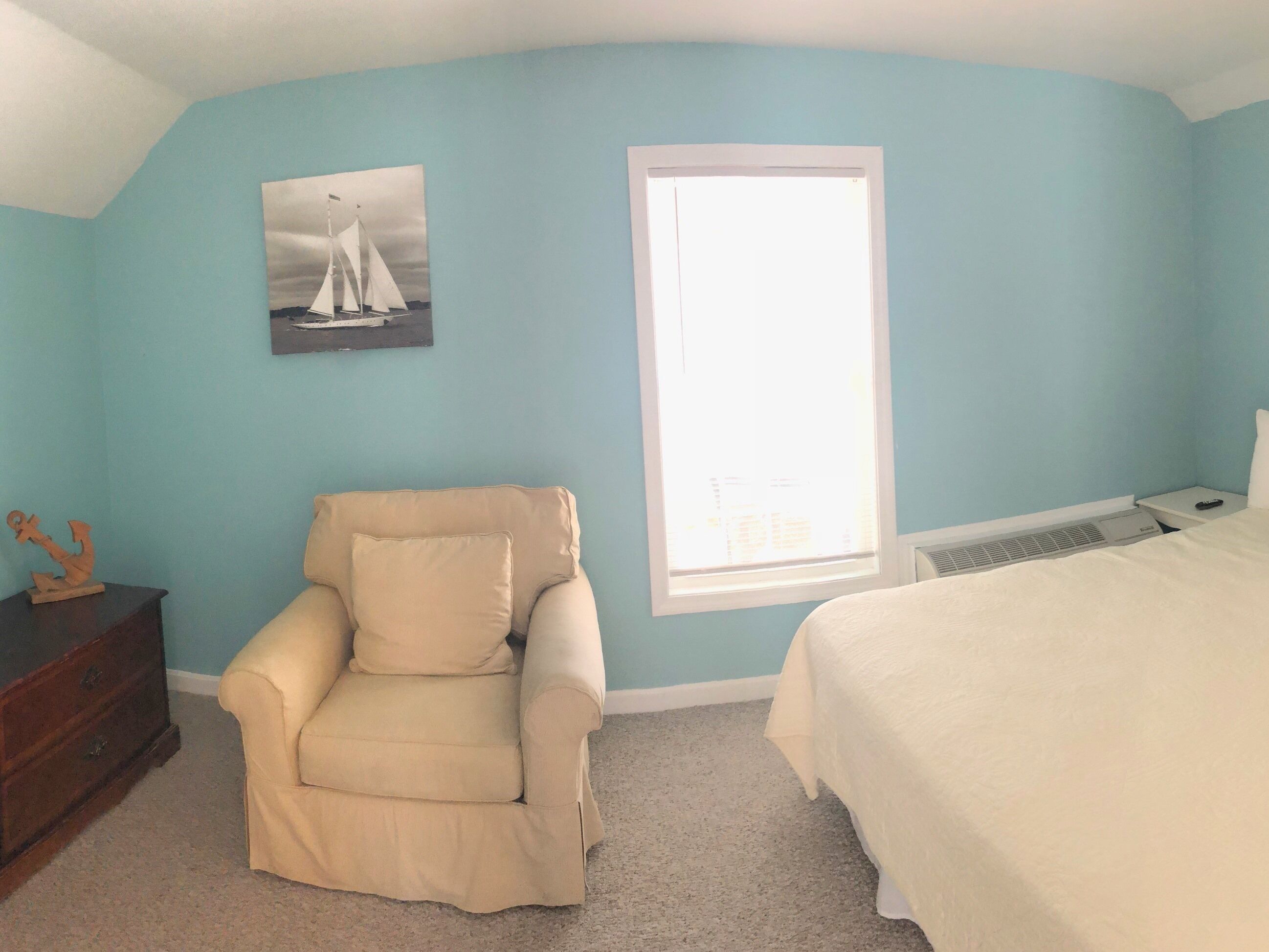 A bedroom with a teal wall, a beige armchair, a bed with white bedding, a nightstand with a remote control, and a window with blinds.