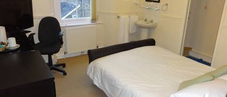 A hotel room with a bed, a desk with a computer, a chair, a TV, a sink, and a window with a view of buildings.