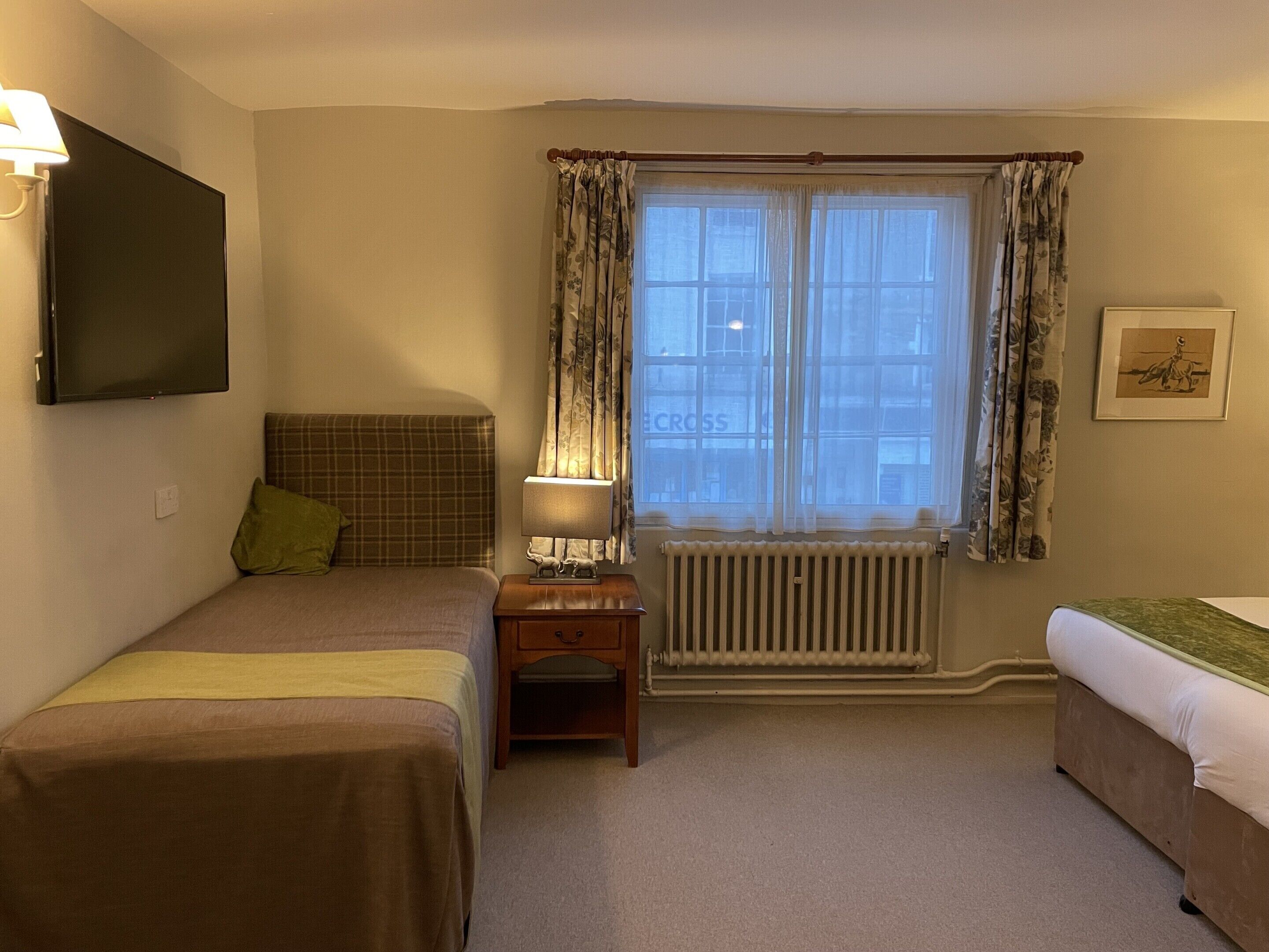 A hotel room with two beds, a small table, a lamp, a radiator, and a window with curtains.
