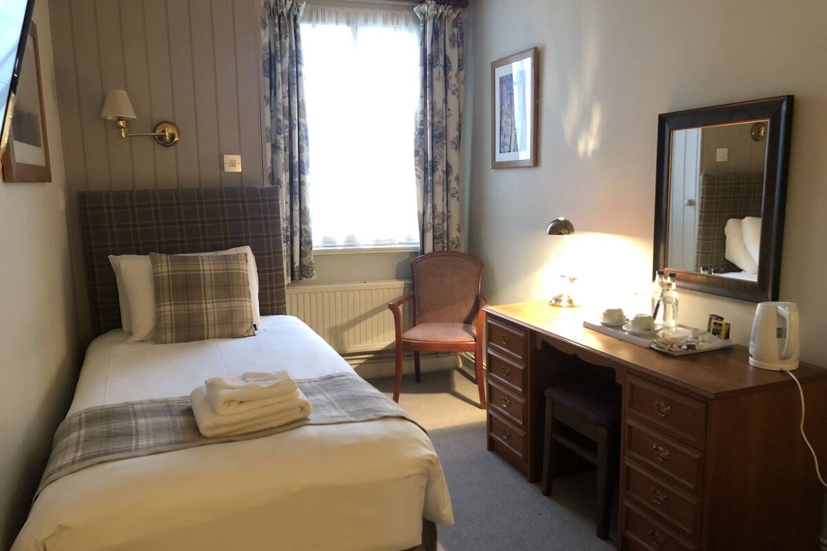 A hotel room with a bed, a desk, a chair, a lamp, a mirror, and a radiator.