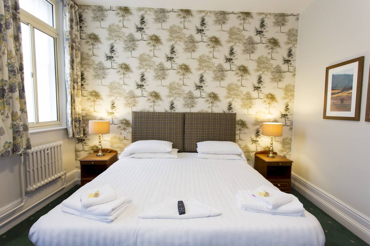 A double bed with white linens, two bedside tables with lamps, a radiator, and a patterned wallpaper.