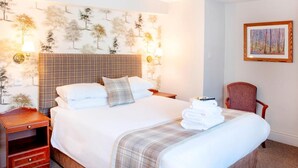 A neatly made bed with a plaid headboard, a bedside table with a lamp, and a chair. - The Old Bell Inn (Warminster)