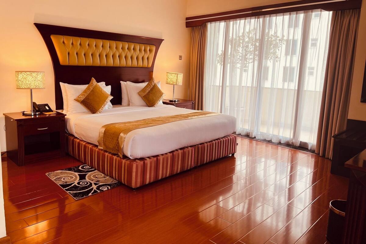 A hotel room with a large bed, two bedside tables with lamps, a striped bench, and a large window with curtains.