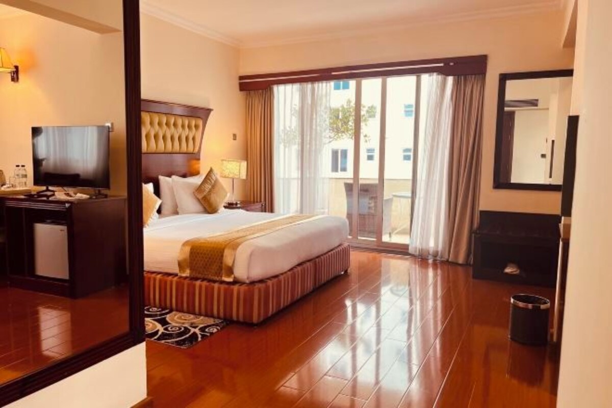 A hotel room with a bed, a television, a desk, a chair, a trash bin, and a large window with curtains.