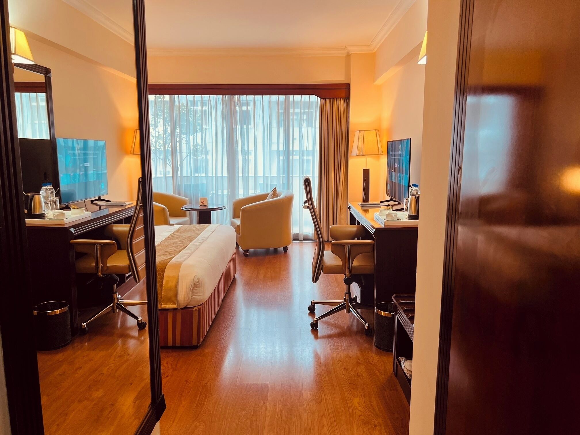 A hotel room with a bed, two armchairs, a desk with a computer, and a large window with curtains.