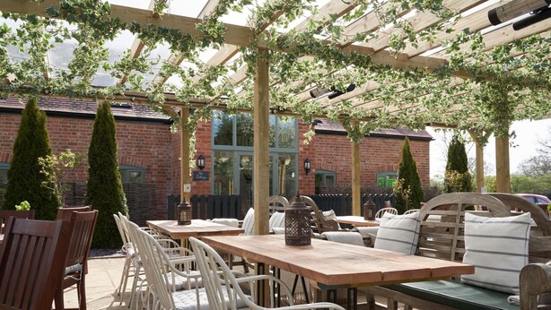 Une terrasse extérieure avec des tables et des chaises en bois, une pergola couverte de plantes grimpantes et, au fond, des bâtiments en briques.