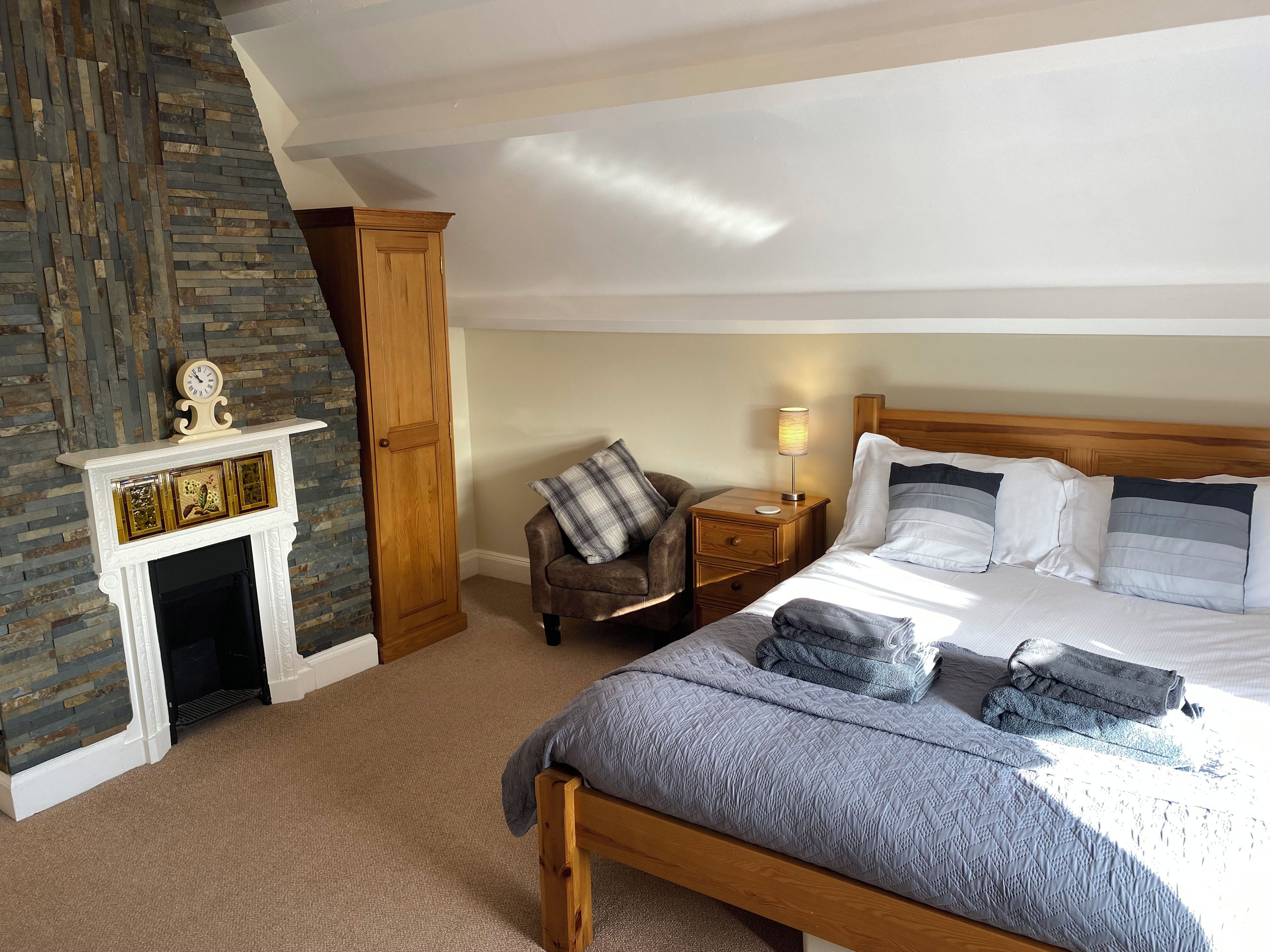 A bedroom with a bed, a nightstand, a chair, a fireplace, and a wooden wardrobe.