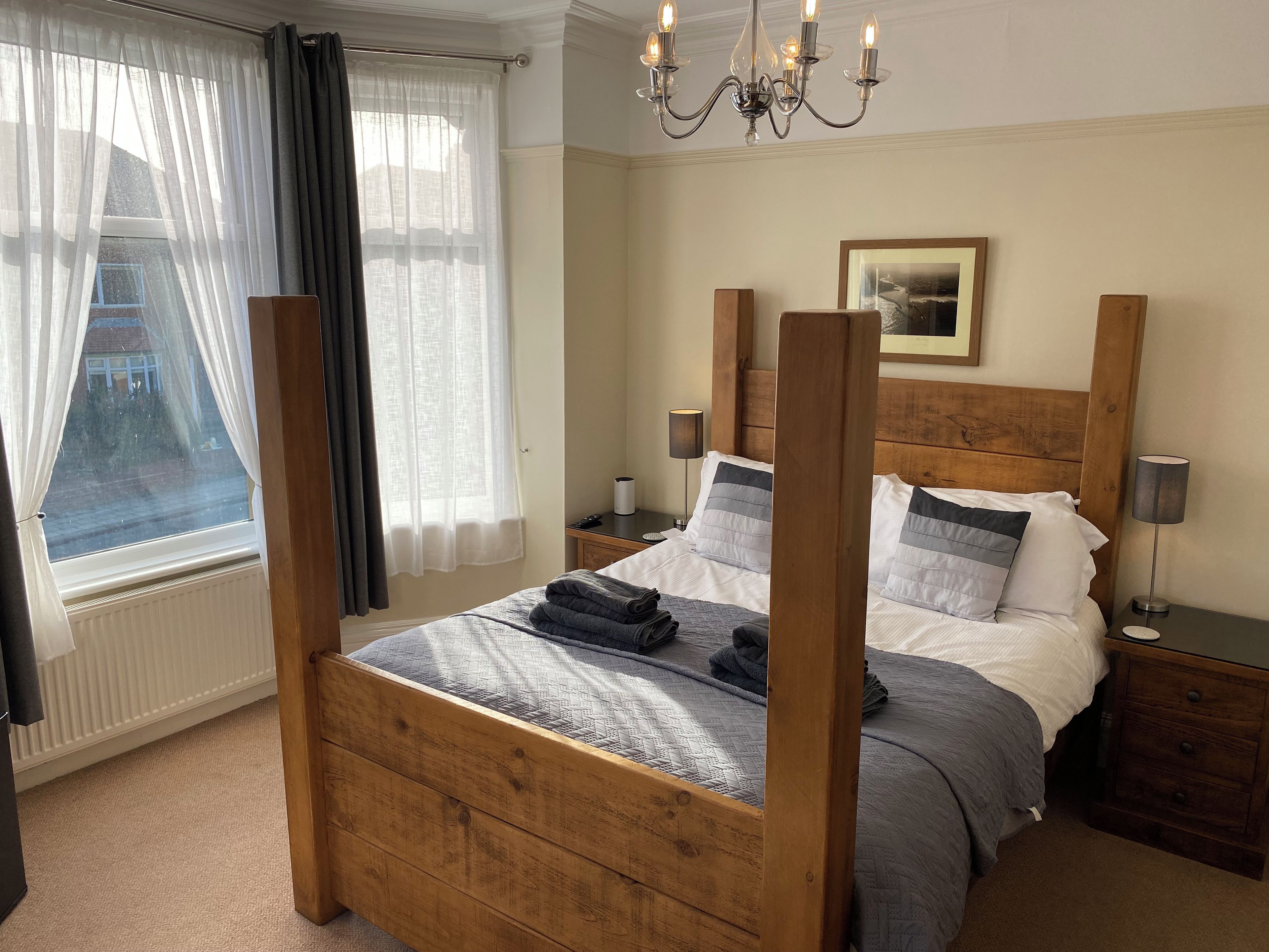 A four-poster bed with a wooden headboard, a bedside table with a lamp, and a view of the outside through a window with sheer curtains.