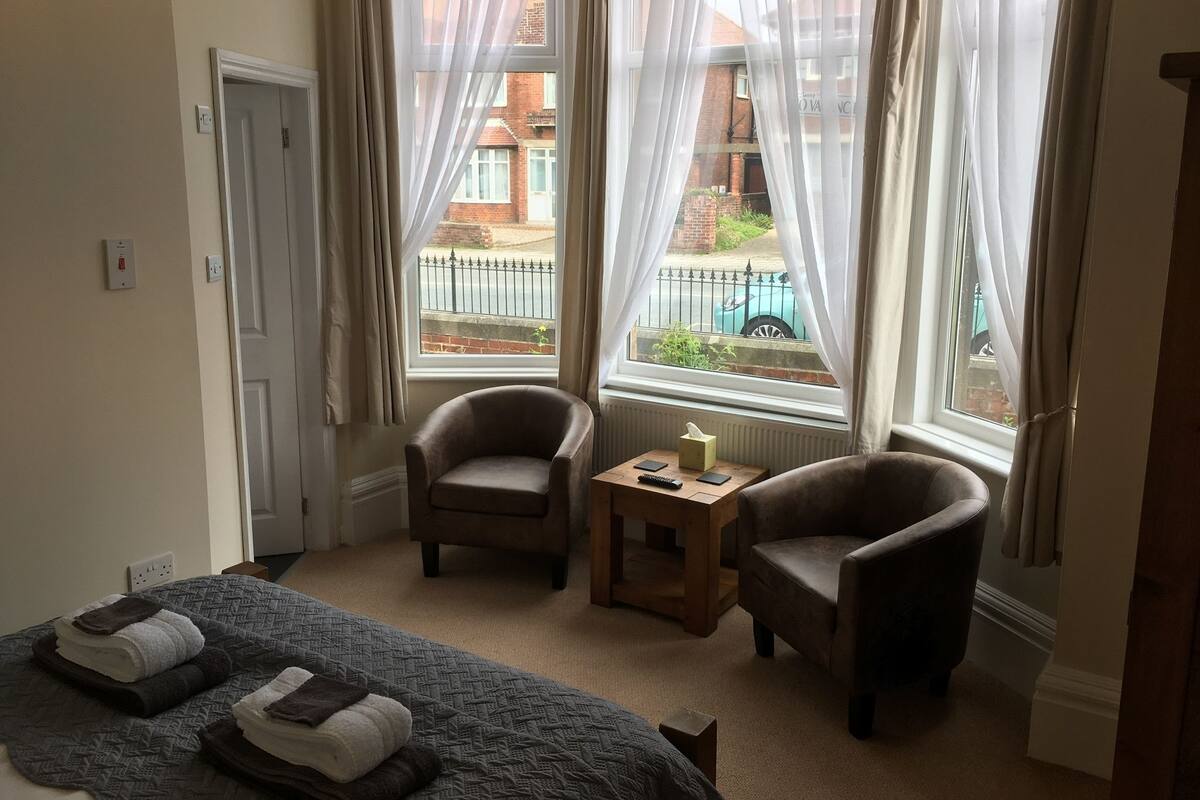 Habitación de hotel con cama, dos sillones, una mesita y vistas a la calle a través de amplios ventanales.