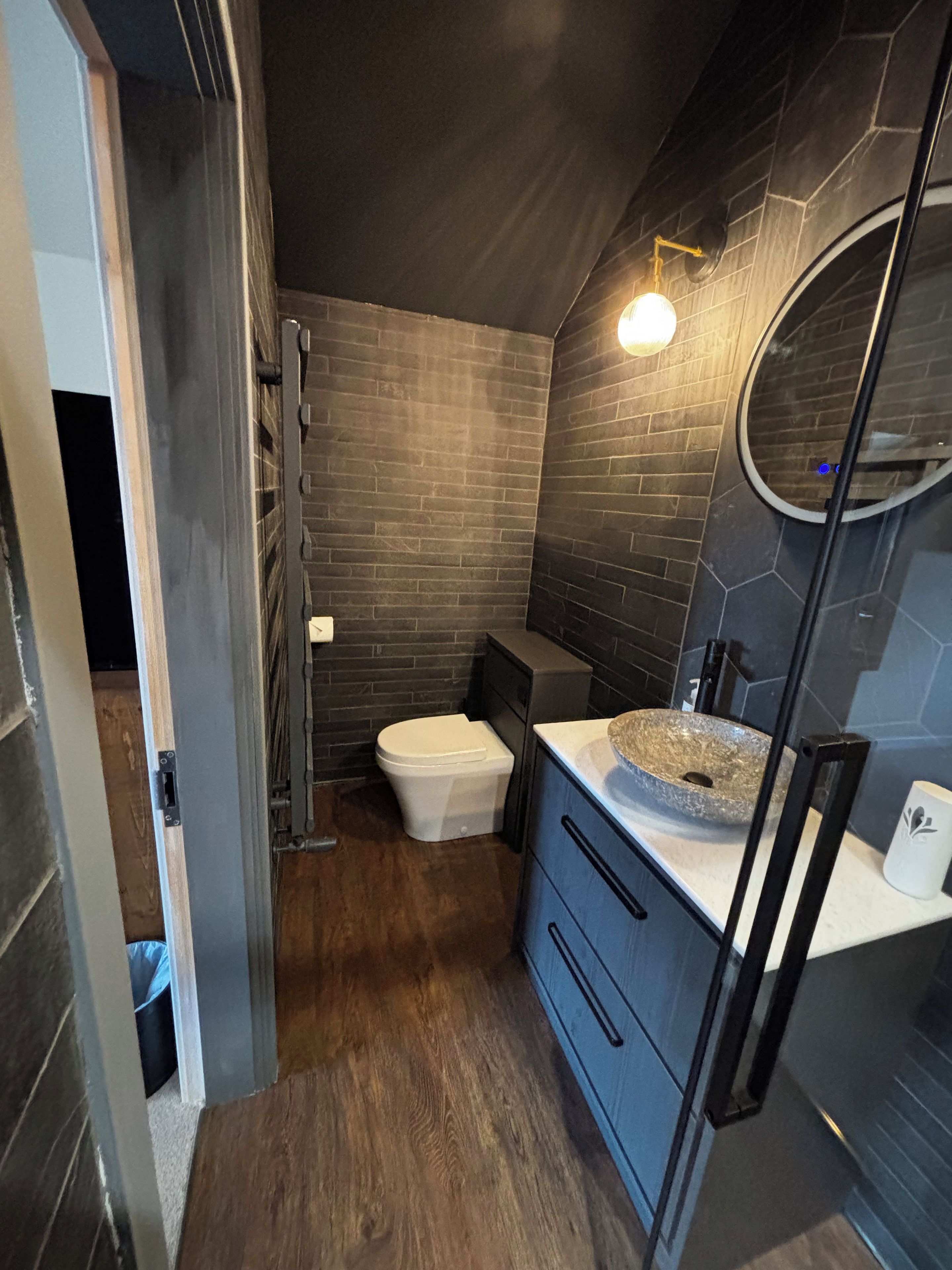 A modern bathroom with a stone sink, dark tiled walls, and a wooden floor.