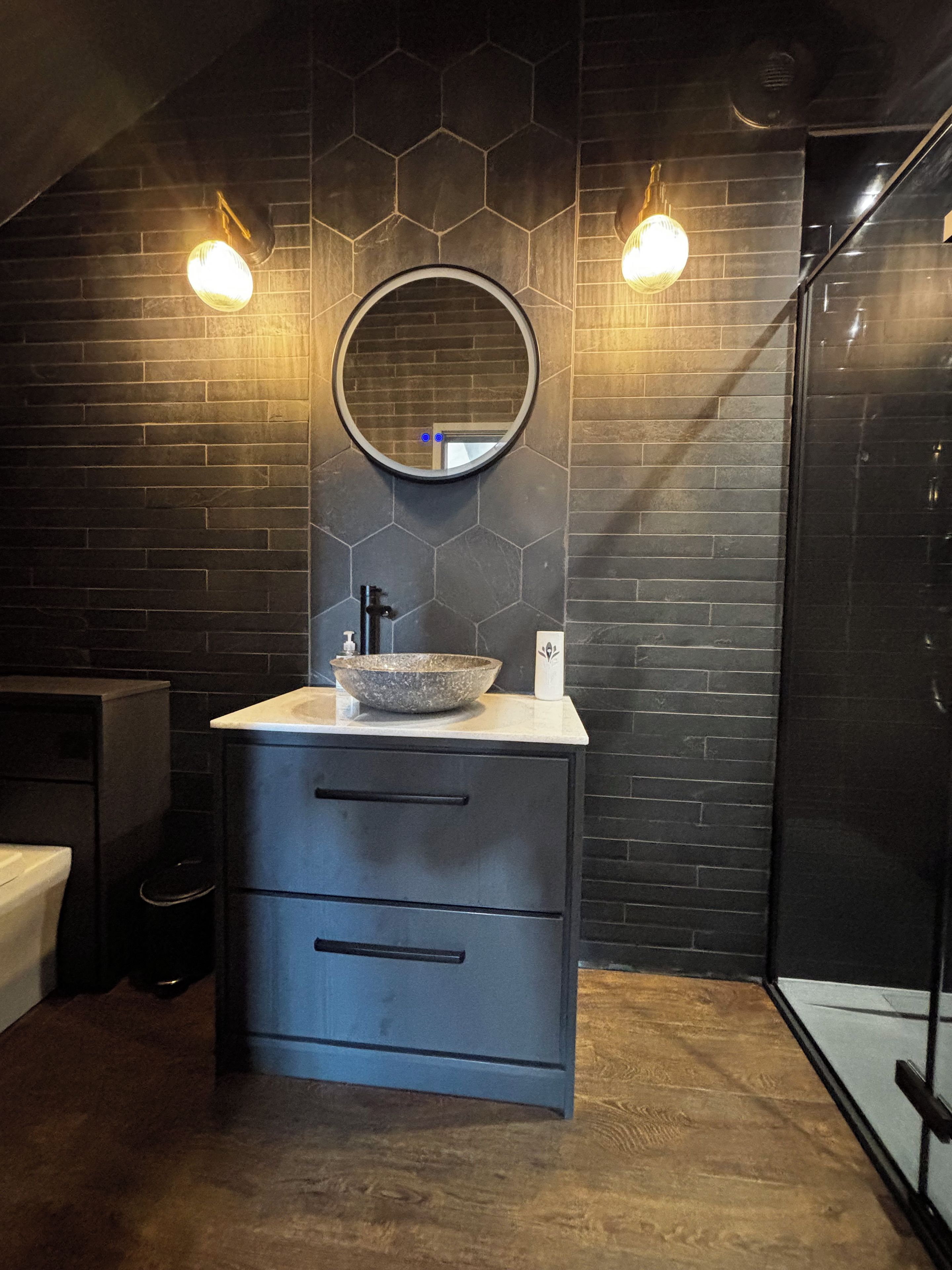 A bathroom with a round mirror, a vessel sink, and a dark tiled wall.