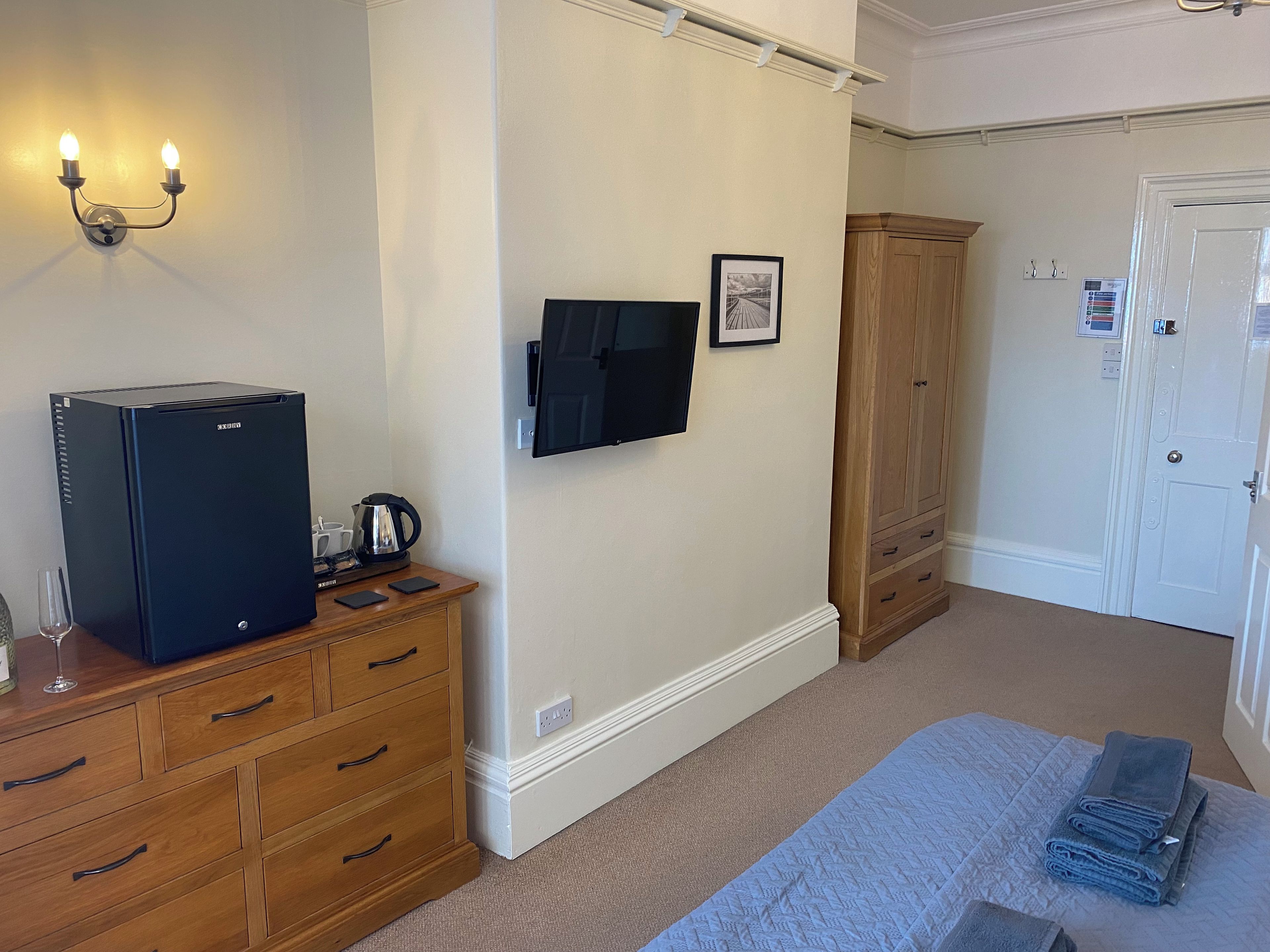 A hotel room with a bed, a wooden dresser, a black refrigerator, a wall-mounted television, and a wooden wardrobe.