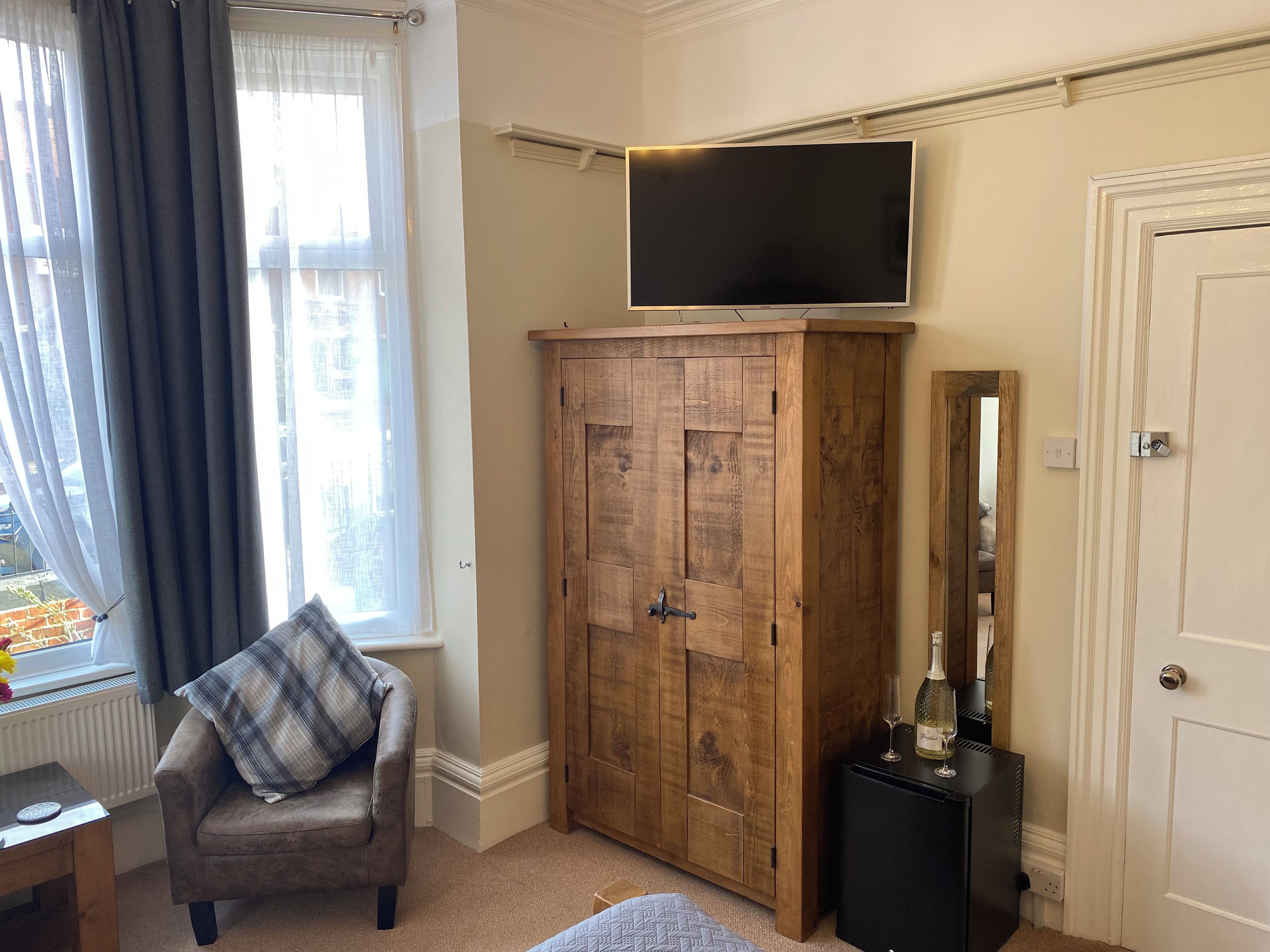 Quarto com televisão na parede, guarda-roupa de madeira, cadeira com almofada xadrez e uma mesa pequena com uma garrafa e copos.