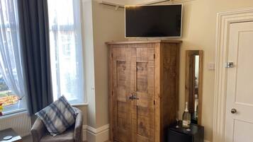 Quarto com televisão na parede, guarda-roupa de madeira, cadeira com almofada xadrez e uma mesa pequena com uma garrafa e copos.