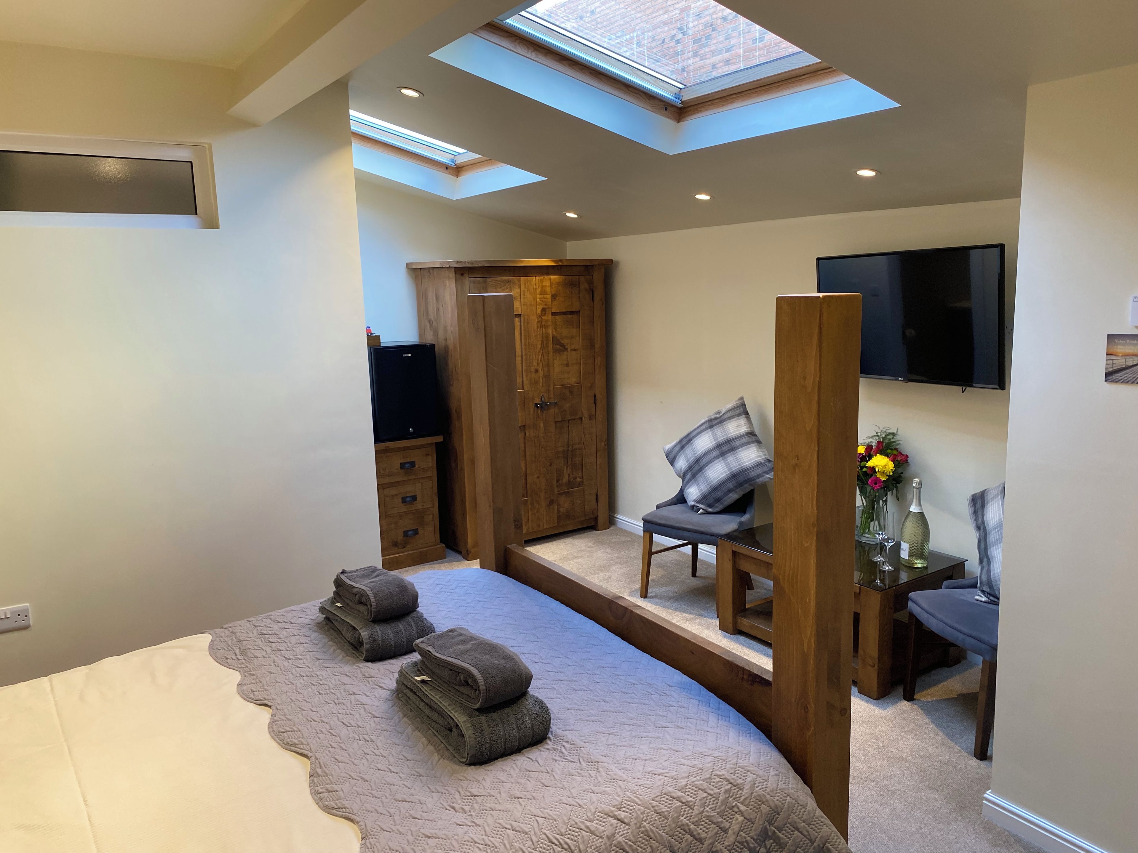 Een slaapkamer met dakraam, een bed met beigekleurige deken, een houten garderobe, een televisie en een klein tafeltje met bloemen.