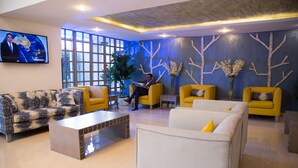 Un vestíbulo moderno con un televisor grande, sofás amarillos y blancos, una mesa de centro y árboles decorativos en la pared.