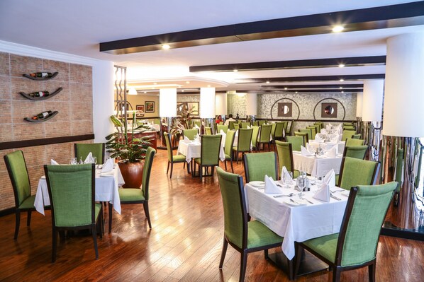 Un restaurant avec des chaises à revêtement vert, des nappes blanches et un mur en briques orné de bouteilles de vin.