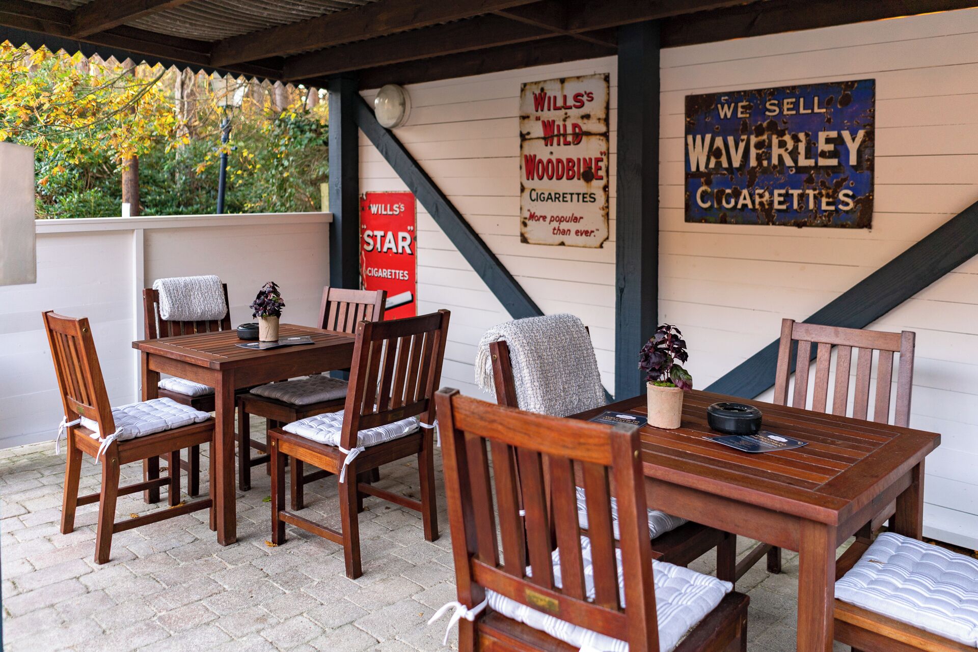 Un espace extérieur aménagé avec des tables et des chaises en bois, un sol en briques et des panneaux publicitaires pour les cigarettes Waverley.