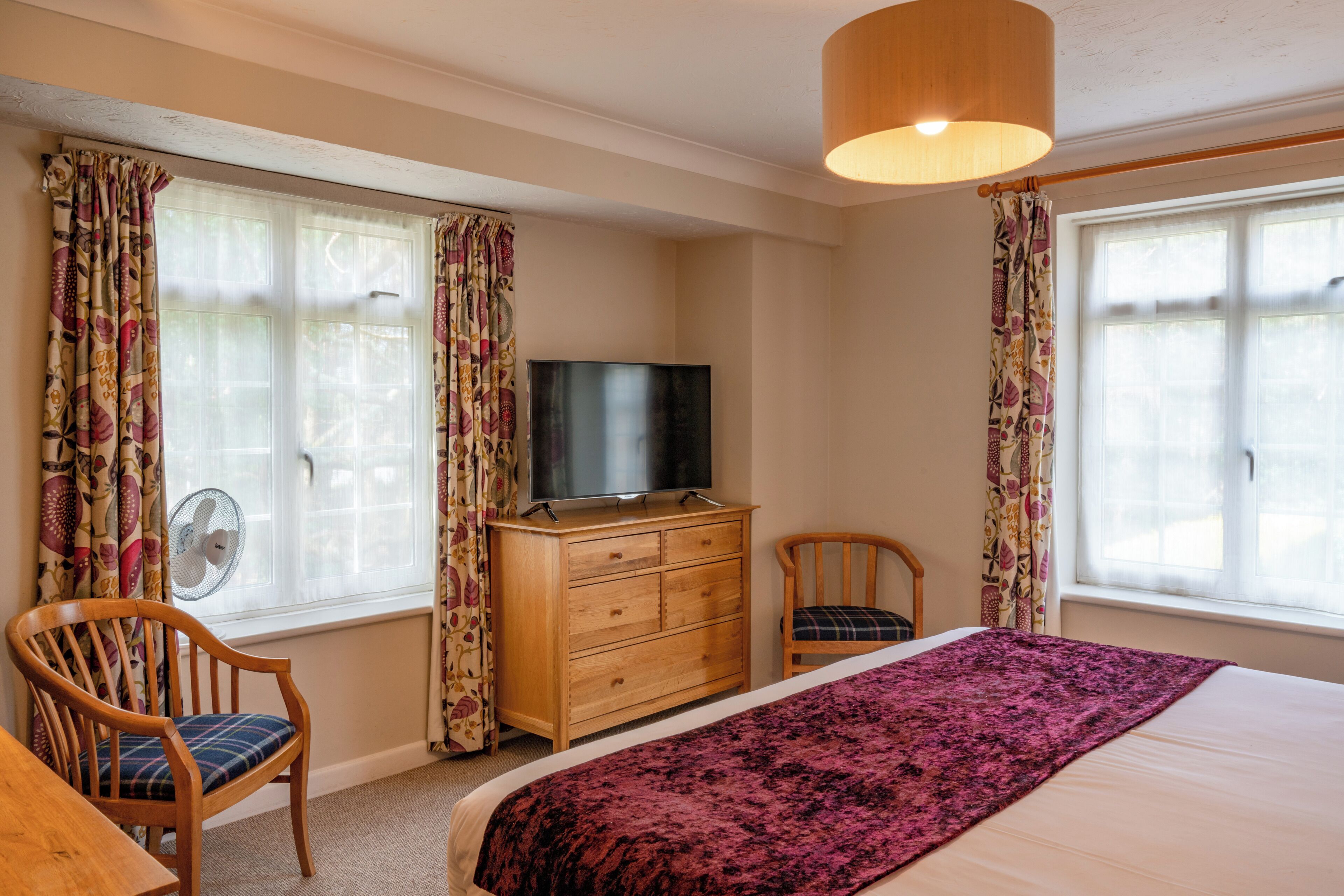 A bedroom with a bed, a wooden dresser, a television, two chairs, and a window with curtains.