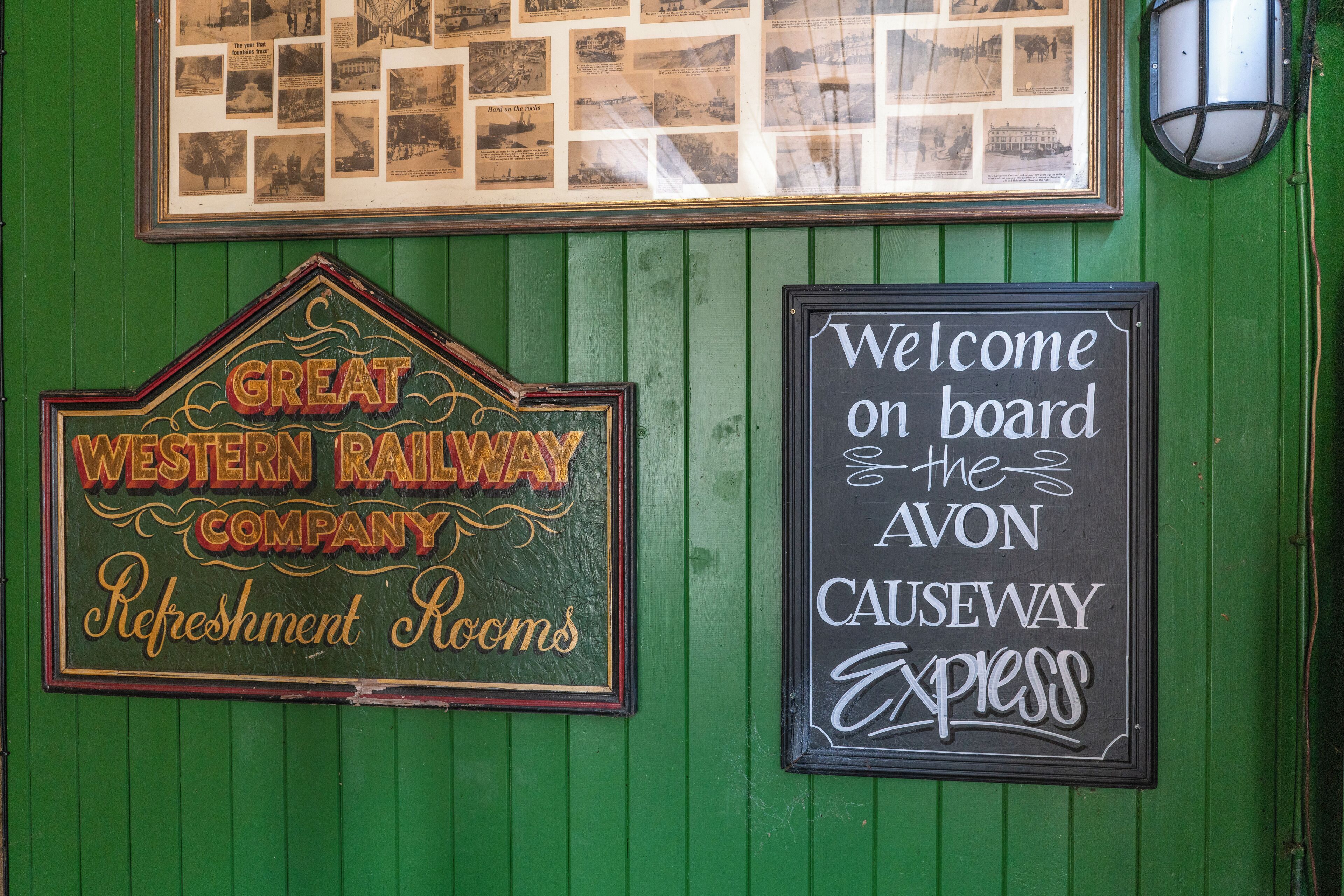 A green wall with two signs: one for the Great Western Railway Company Refreshment Rooms and another welcoming visitors to the Avon Causeway Express.